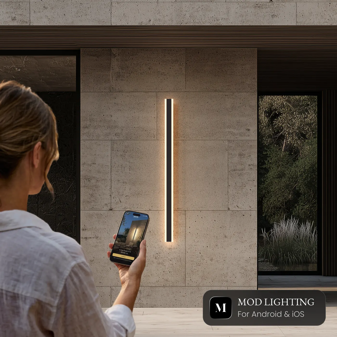 A woman holding her phone with the MOD LIGHTING app open, a Haylen smart wall light lit on the wall in front of her